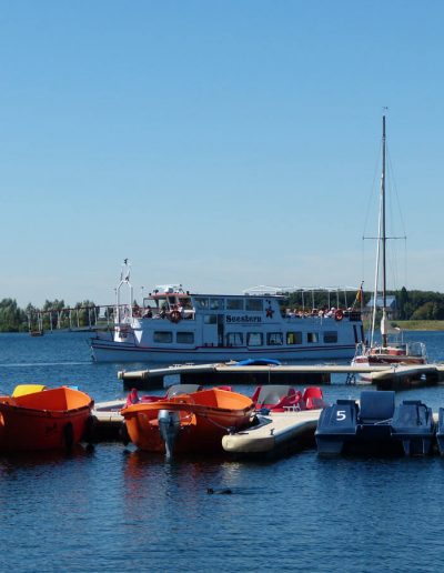 Das Foto zeigt Elektro- und Tretboote, die im Hafen liegen. Im Hintergrund befindet sich das Fahrgastschiff Seestern, das auf der Nord- und Südsee seine Runden dreht.