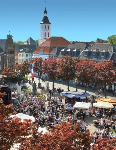 Das Foto zeigt den belebten Marktplatz bei schönem Wetter. Die Menschen feiern ein Fest. Im Hintergrund erhebt sich hinter einer Baumreihe die kleine evangelische Barockkirche.